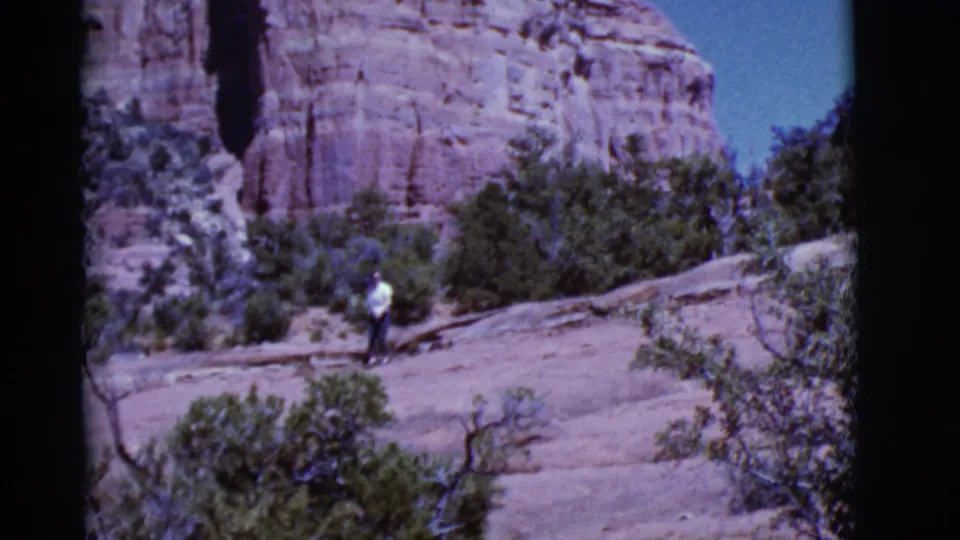 1968 Arizona home-movie frame of a woman walking along pale stone terrain with scrub vegetation and red cliffs behind her.