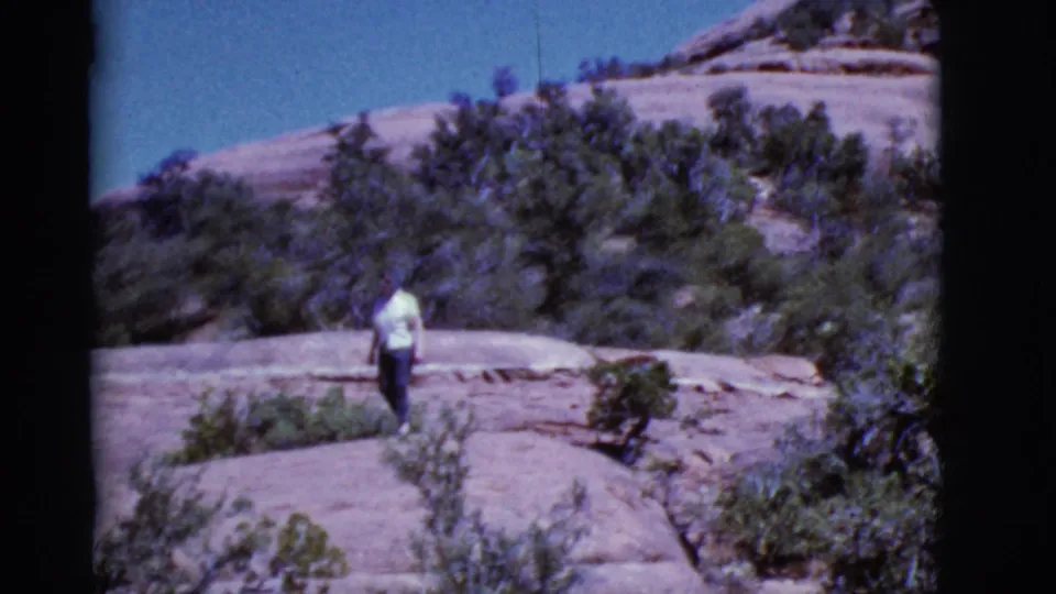1968 Arizona 8mm frame showing the woman from behind as she walks away across flat rock among desert shrubs.
