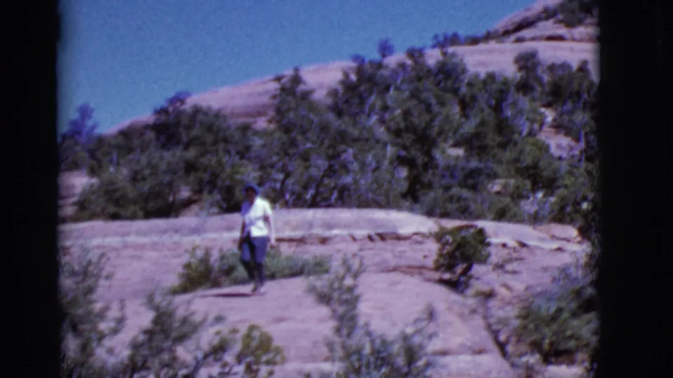 1968 Arizona home-movie frame of a woman in a white top and blue pants walking toward the camera through rocky desert terrain.
