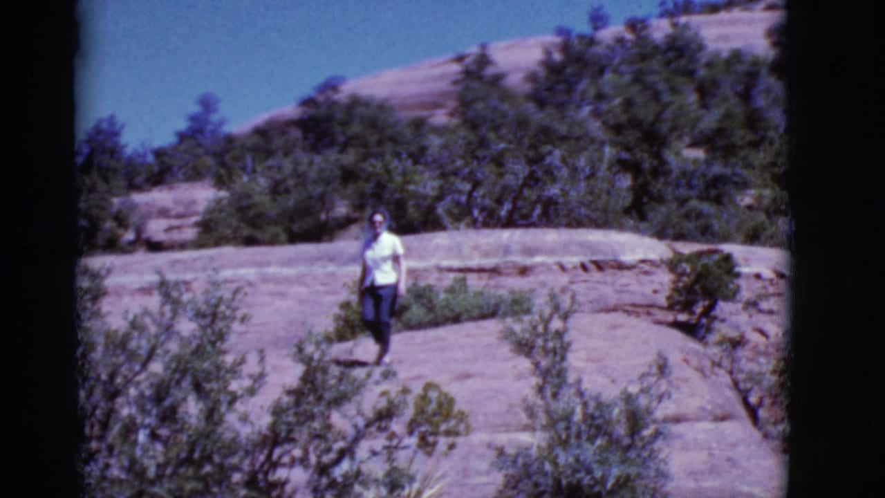 A woman in a light shirt and dark pants walks across rocky Arizona desert terrain in 1968 home-movie footage.