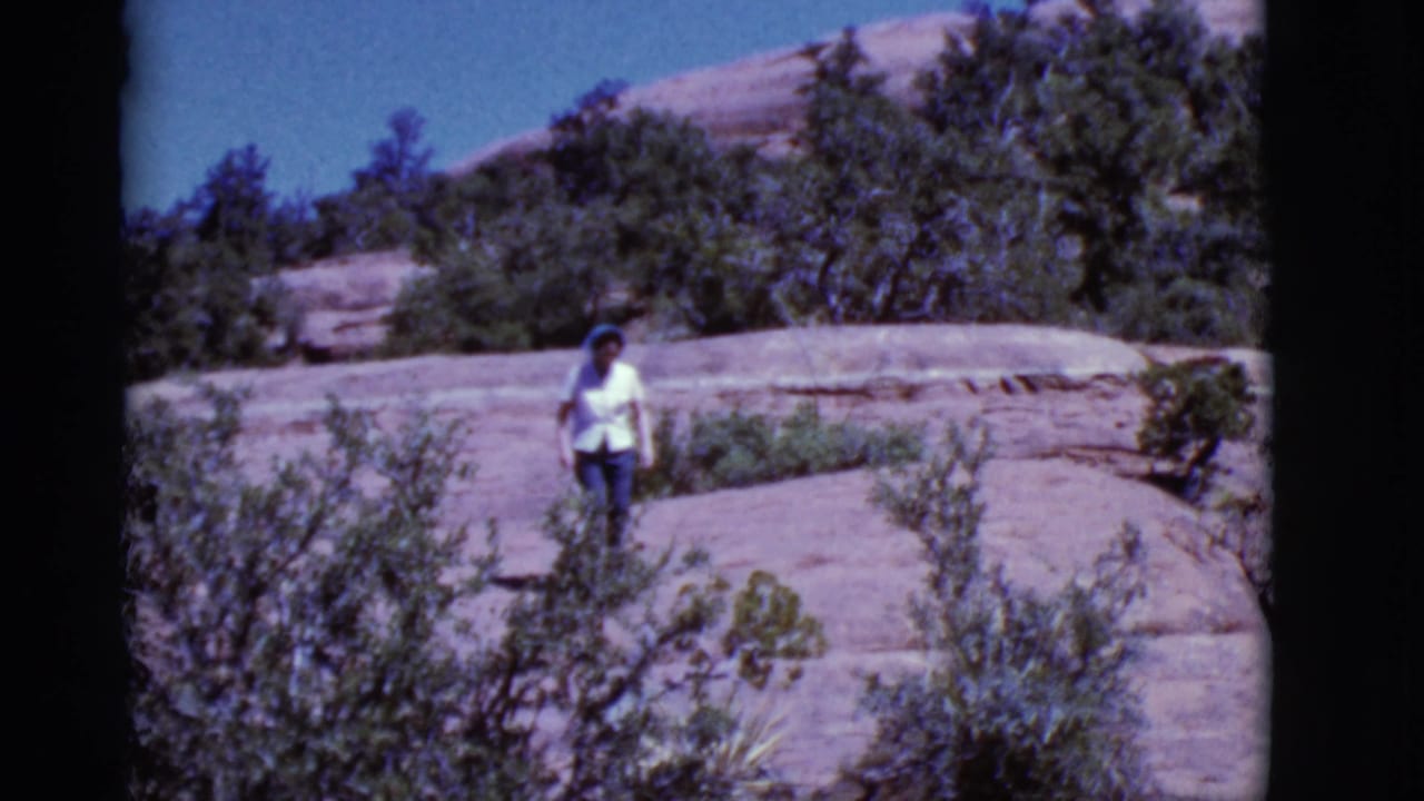 Woman farther across a rocky slope with shrubs and blue sky visible.
