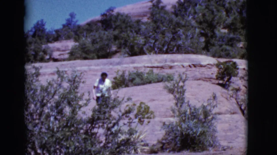 A woman moves through low desert shrubs on layered Arizona rock in a handheld 1968 home-movie frame.