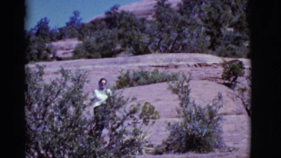 The hiker appears near foreground brush with layered Arizona rock and trees behind her in 1968 8mm footage.
