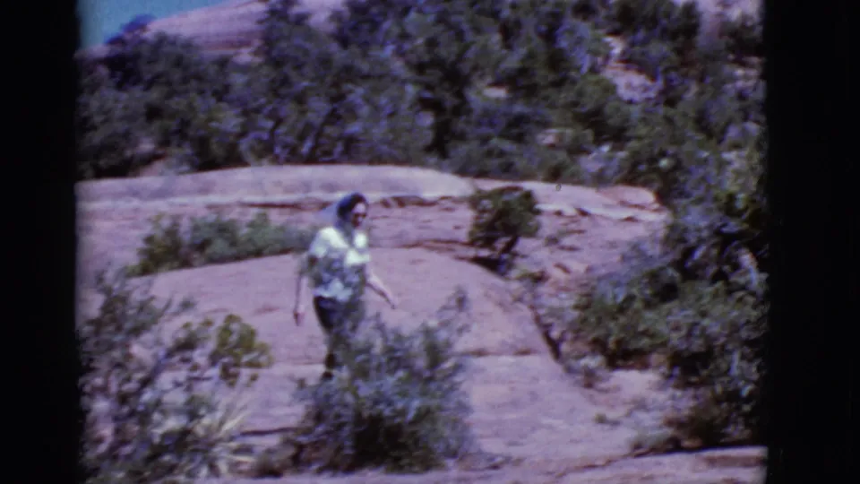 The woman continues along a sunlit rock path in the likely Arizona desert, with scrub plants and red formations around her.