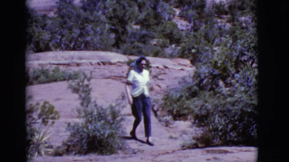 A woman in a white shirt and dark pants walks closer to the camera on a desert rock path, with shrubs in the foreground.