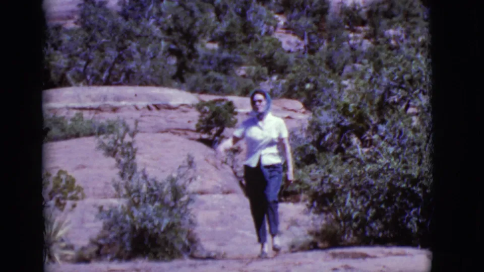 Near the end of the clip, the woman faces the camera on the sunlit rock trail, surrounded by sparse shrubs and trees.