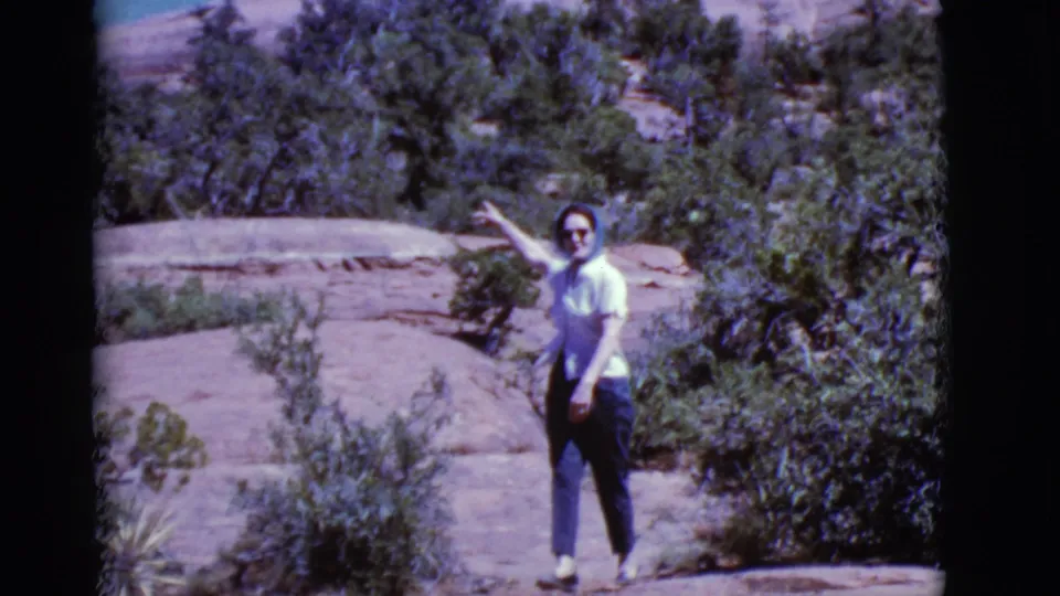 In 1968 Arizona home-movie footage, a woman in sunglasses and a blue headscarf points across a rocky desert landscape.