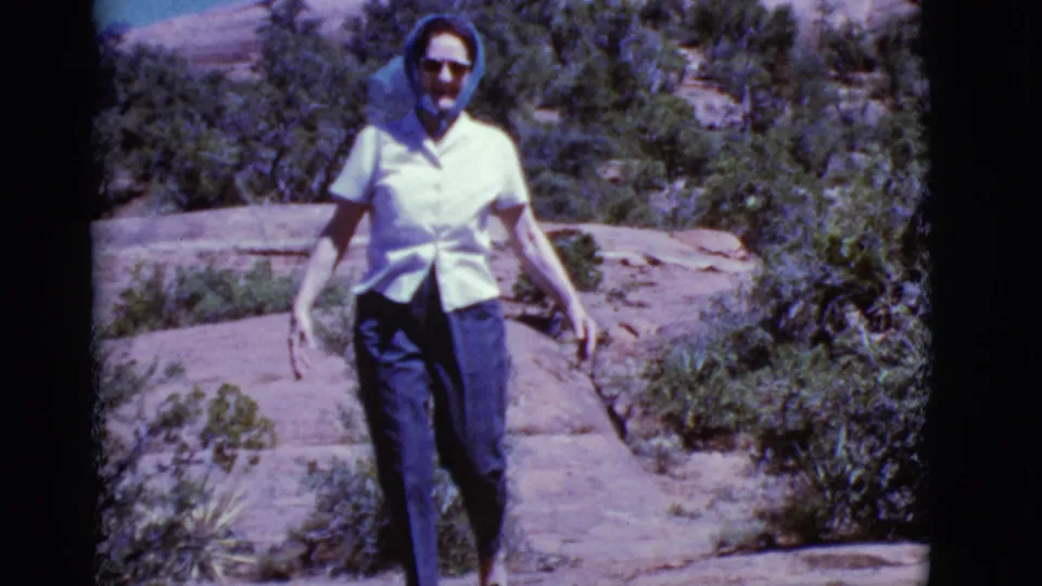 Closer 1968 Arizona frame of a woman in sunglasses and a blue headscarf walking through a rocky desert landscape.