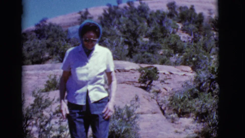 The woman walks near the camera in an Arizona desert scene, with sunglasses, headscarf, rocky ground, and scrub vegetation visible.