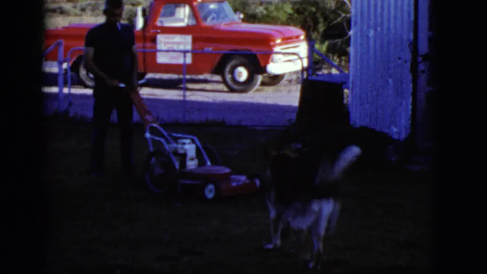 A man stands with a lawn mower in a shaded yard while a red pickup and blue shed appear in the background.