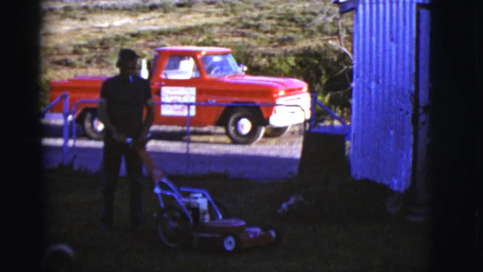 1968 Arizona home-movie frame with the man standing over the red mower in deep shade, a German Shepherd partly visible, and a red pickup beyond.