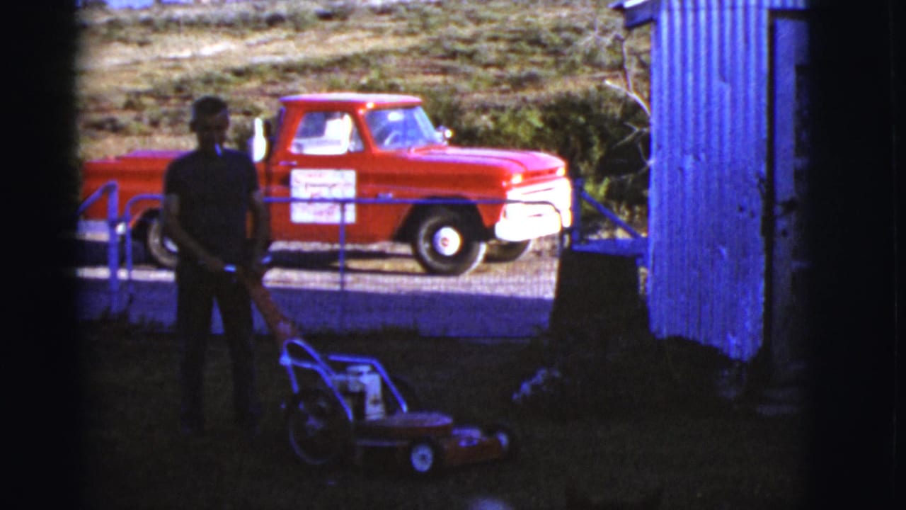 Man stands with a push mower in a shaded yard, with a red pickup behind a fence.