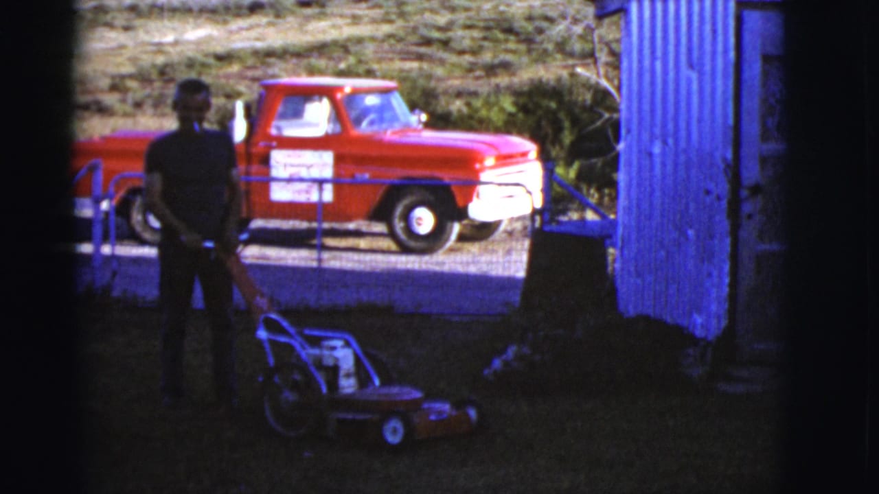 Man holds a mower in a dark yard area with a red pickup visible outside the fence.