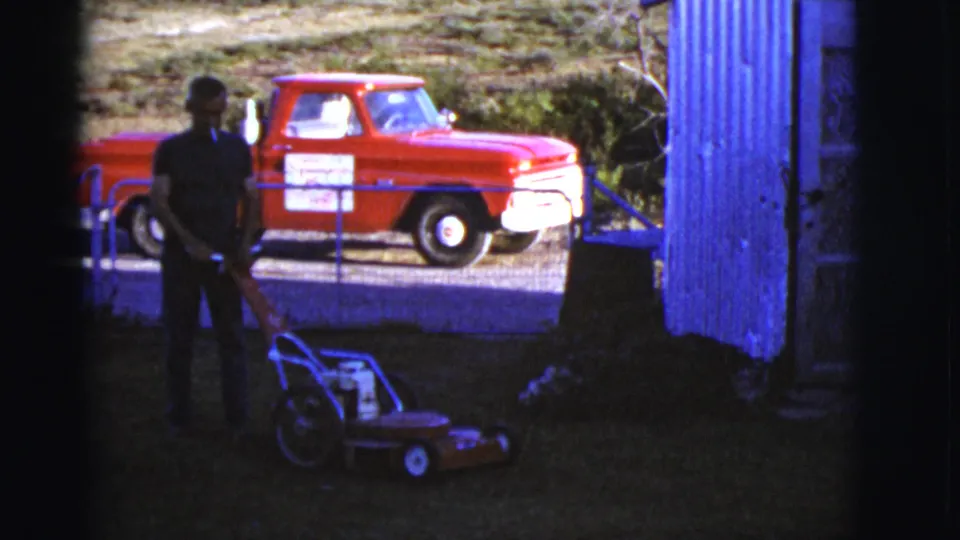 1968 Arizona yard scene with the man and mower in shade, a red pickup beyond the fence, and corrugated siding at right.
