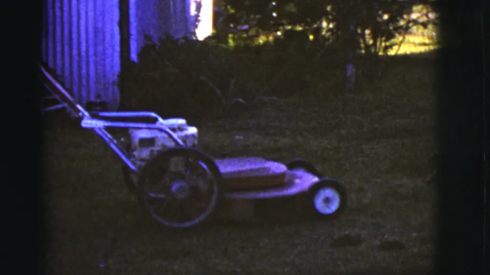 A red walk-behind lawnmower sits on grass in a shadowed backyard, framed by dark 8mm edges and nearby shrubs.