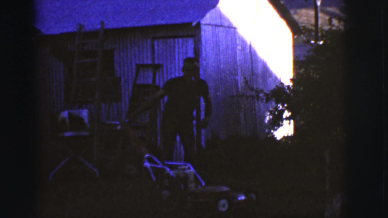 1968 Arizona home-movie frame with an adult man silhouetted near a push lawnmower and wooden shed in a shadowed backyard.