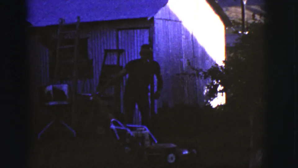 1968 Arizona home-movie frame with an adult man silhouetted near a push lawnmower and wooden shed in a shadowed backyard.