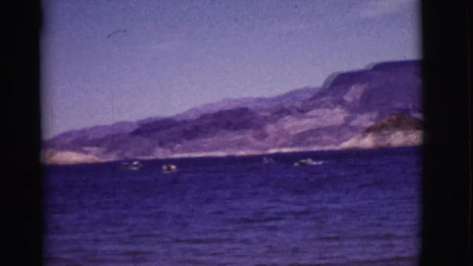 1968 Arizona home-movie frame showing blue water, tiny boats, a pale desert shoreline, and rugged mountains.