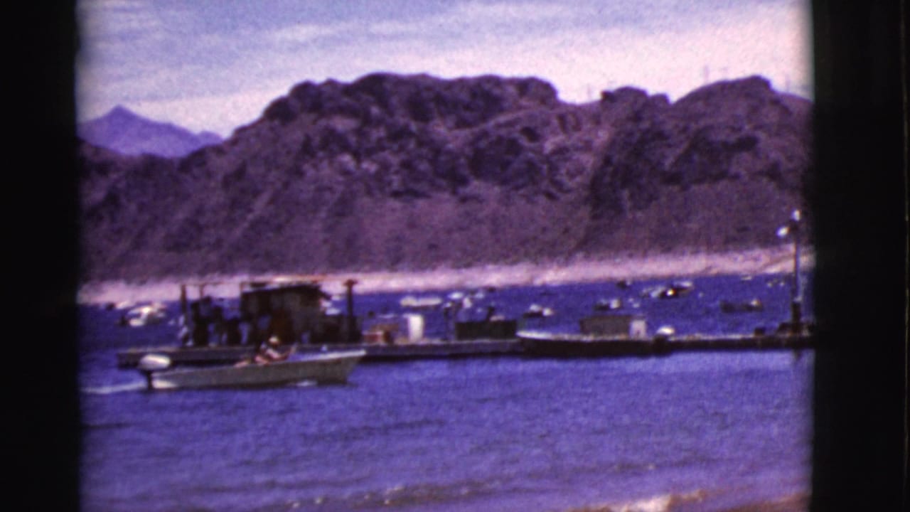 A small motorboat nears the dock in a 1968 Arizona lakeside home-movie frame, with distant people and rocky slopes beyond.