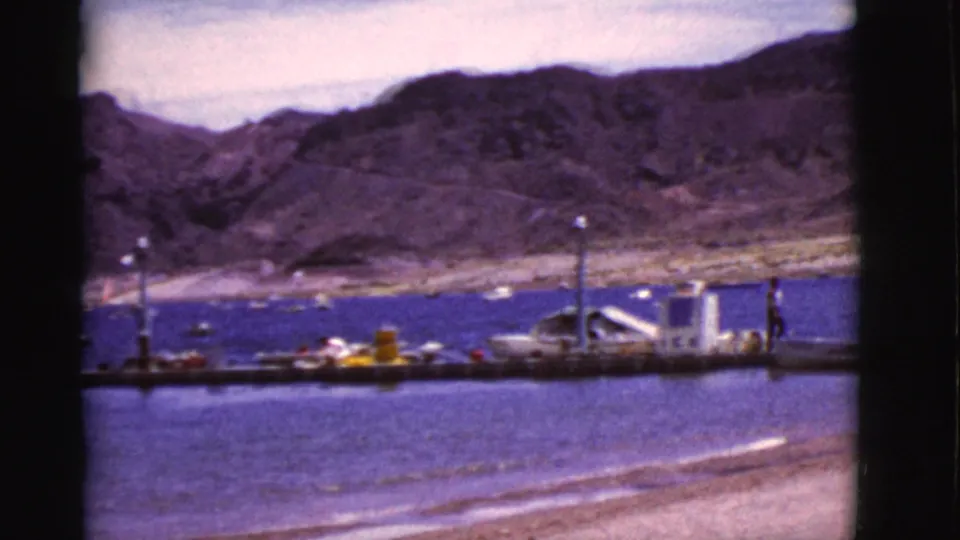 A 1968 Arizona 8mm frame shows a docked boat at a pier, distant people, rippled water, and arid hills beyond the lake.