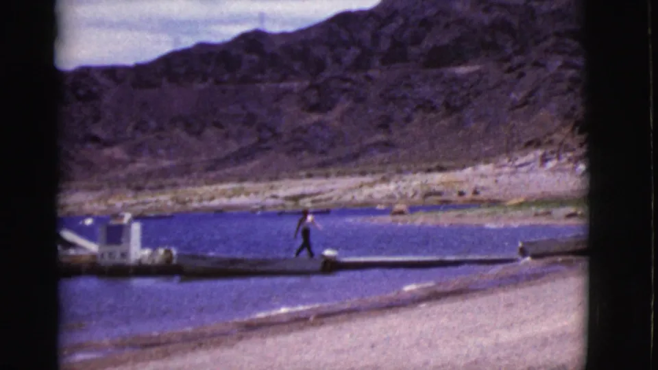 A person walks along a narrow dock extending across blue lake water, with a sandy beach and rocky Arizona hills beyond.