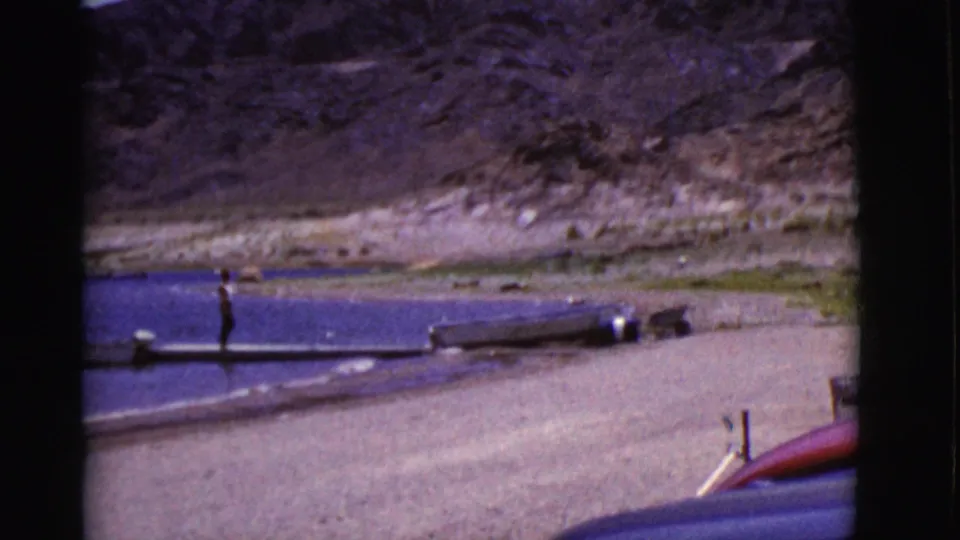 A small figure is seen on the dock near the Arizona lake shoreline, with boats pulled close to shore and mountains in the distance.