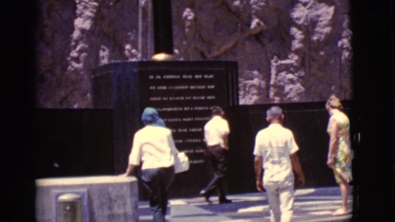 Visitors approach a dark memorial panel in a sunlit stone plaza.