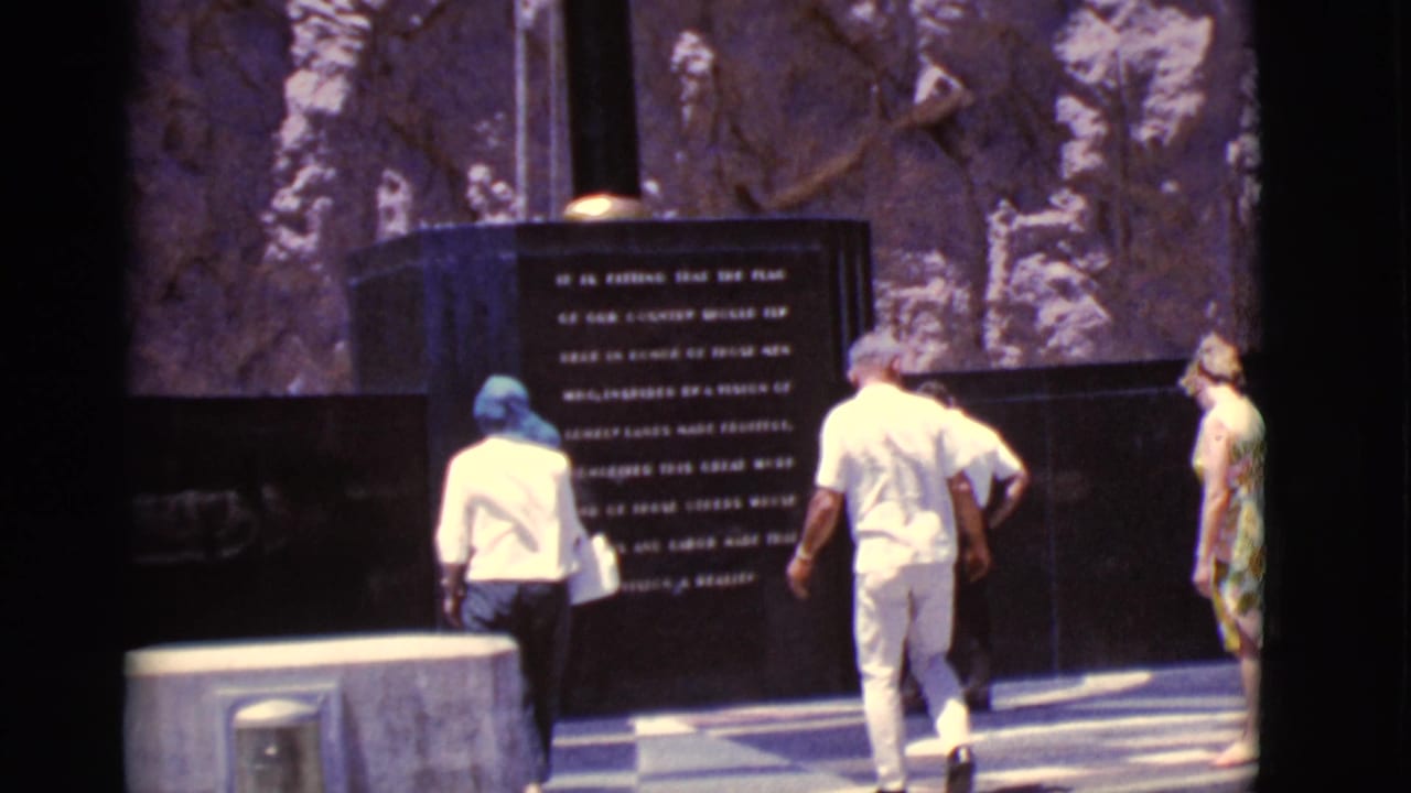 1968 Arizona footage frame with visitors in light clothing approaching and pausing near a dark monument panel.