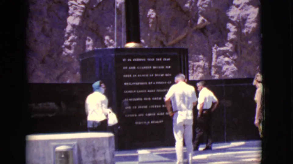 1968 Arizona home-movie frame showing visitors paused respectfully near a memorial, with a rocky backdrop and strong shadows.