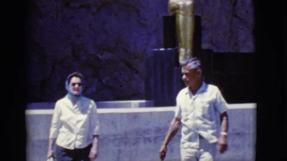 1968 Arizona home-movie frame showing an older couple in light clothing walking past a dark wall and gold-toned statue.
