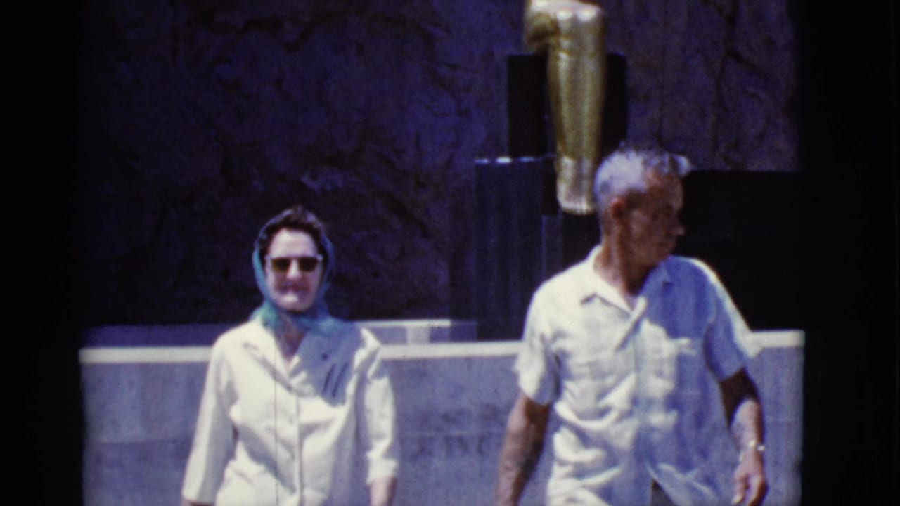 Older woman in sunglasses and older man in a white shirt walking past a statue wall.