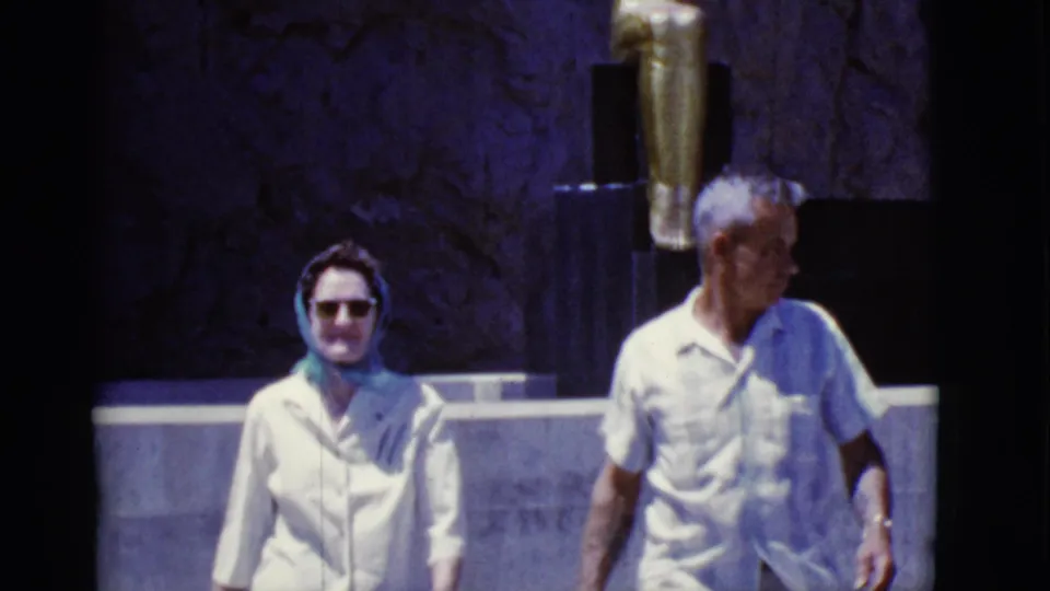 1968 Arizona frame with an older woman in a headscarf and sunglasses walking beside an older man near a monument-like wall.