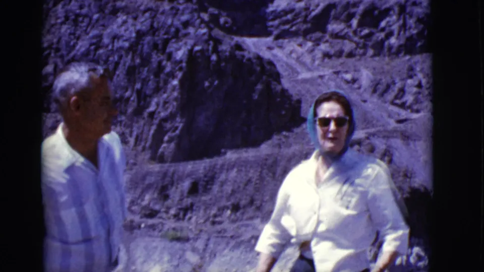 Two adult travelers in 1968 Arizona stand in bright sun on a rocky desert overlook, with a winding road behind them.