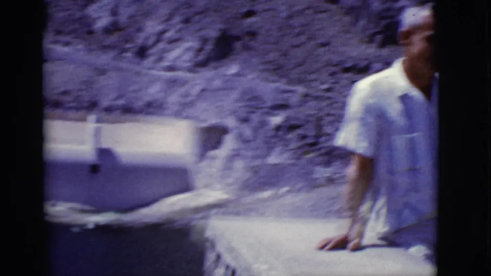 A handheld pan catches the man partly blurred as the view opens to a concrete dam wall and dark water in the Arizona desert setting.
