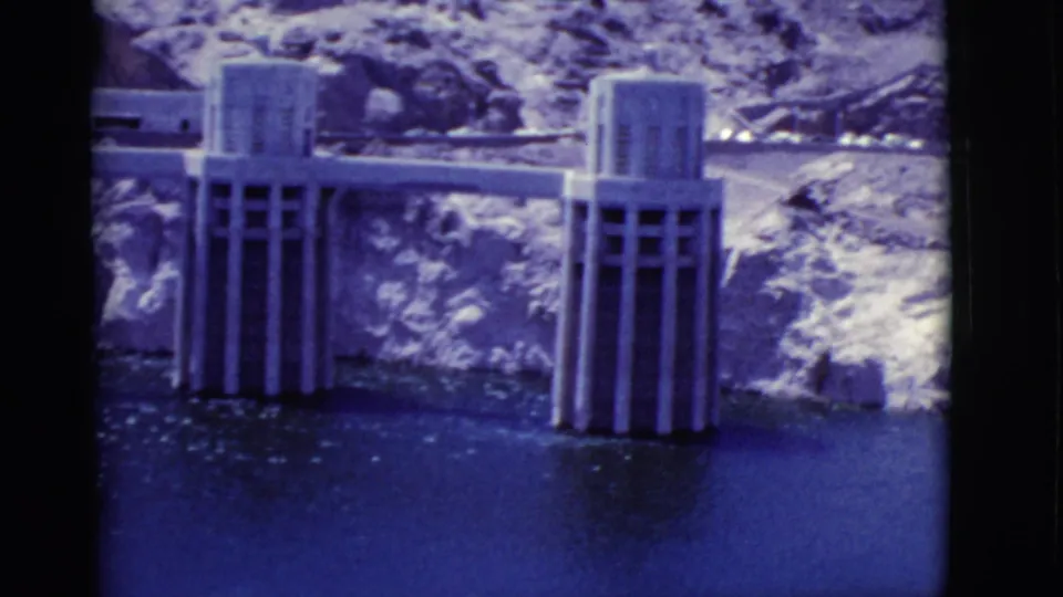 1968 home-movie frame of Hoover Dam intake towers in Lake Mead, with rocky desert slopes and bright water reflections.