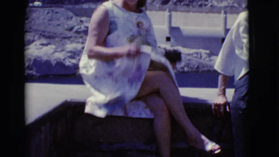 A woman in a light dress sits on a stone ledge at a Hoover Dam overlook, with Lake Mead and dam structures behind her.