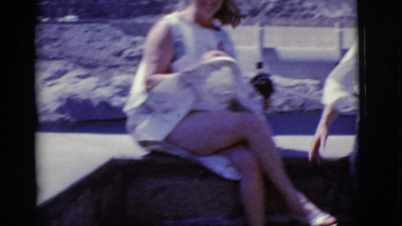 1968 Arizona home-movie frame of a woman in sunglasses and a sleeveless light dress seated near rocky terrain.