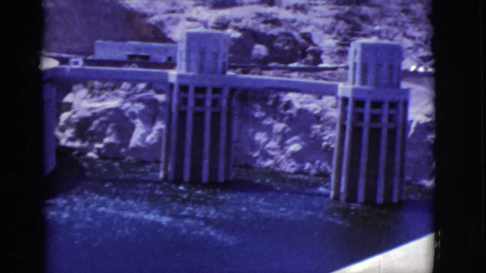 Concrete dam structures stand against rocky Arizona terrain with blue-purple water in aged 8mm footage.
