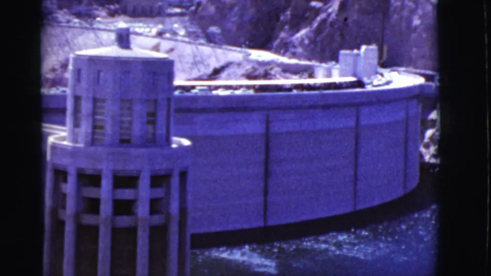 Closer 1968 home-movie frame of a dam model tower and curved wall, tinted purple by aged 8mm film.