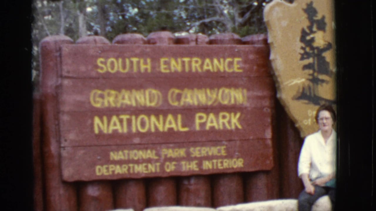 Woman seated beside the Grand Canyon South Entrance sign and National Park Service shield.