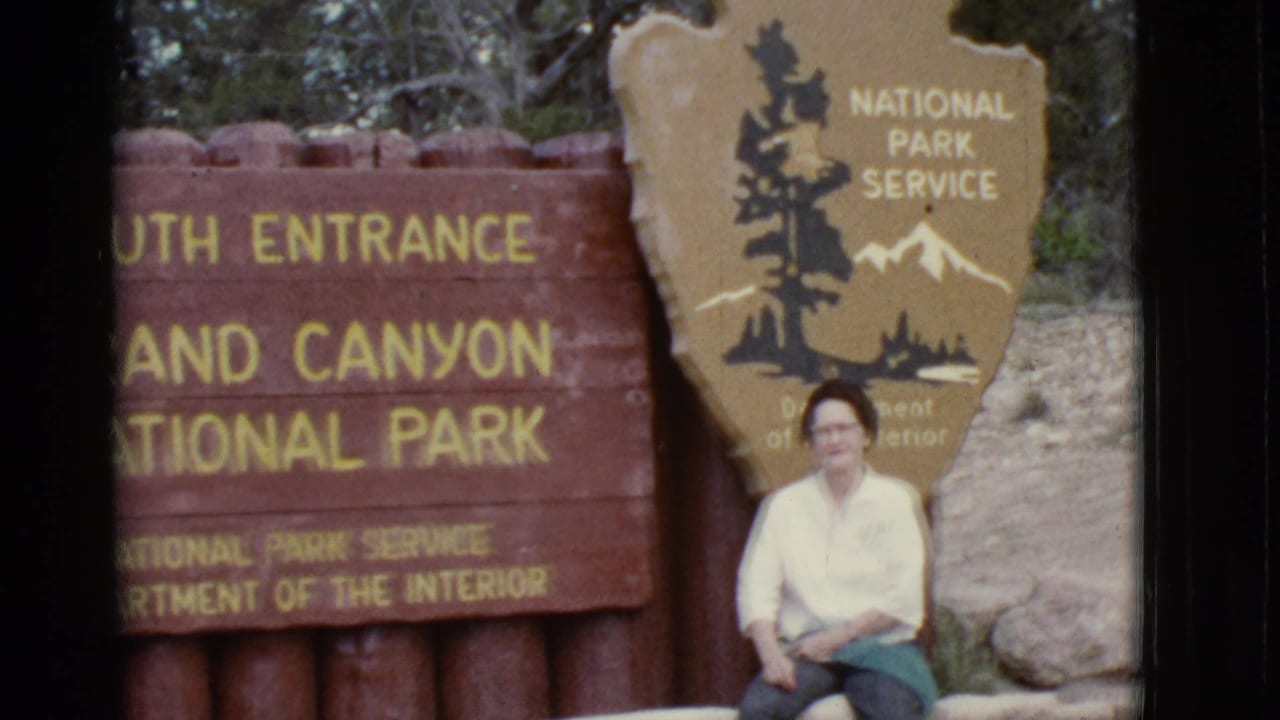 Seated woman in front of the Grand Canyon entrance sign, framed beside the NPS shield.