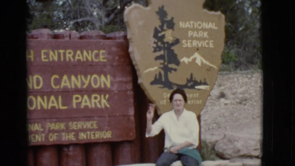 1968 Grand Canyon entrance frame with a seated woman waving near the National Park Service shield.