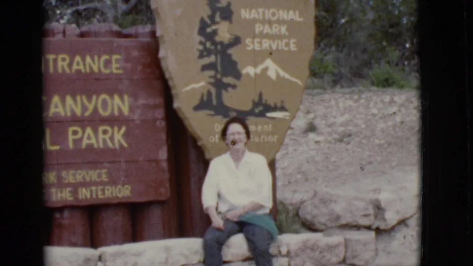 Seated woman at the Grand Canyon National Park South Entrance sign in warm, grainy 8mm footage.