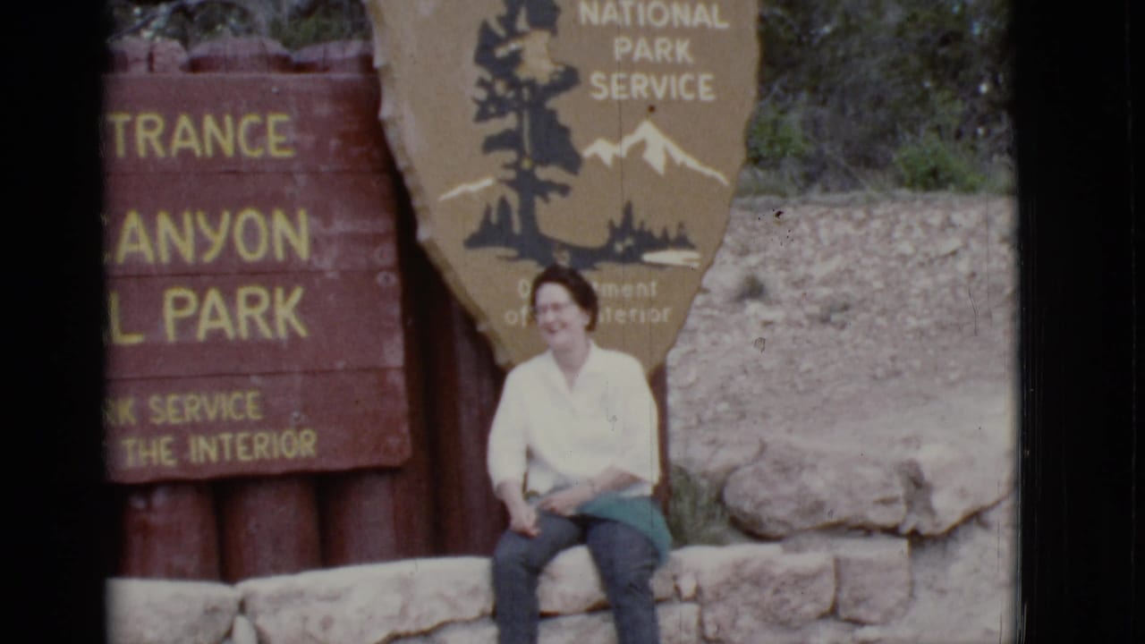 Woman in a white blouse sits on a stone ledge beside National Park Service entrance signs.