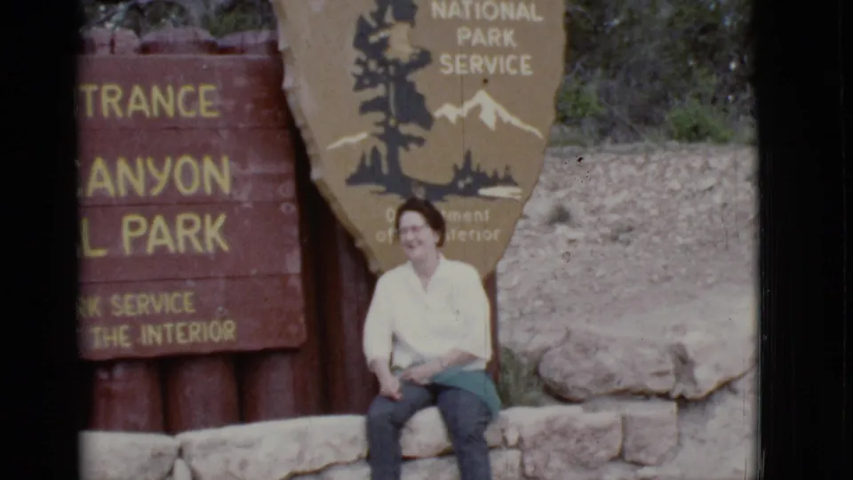 In Arizona 1968 home-movie footage, a woman sits near rustic park entrance signage and looks toward the camera.