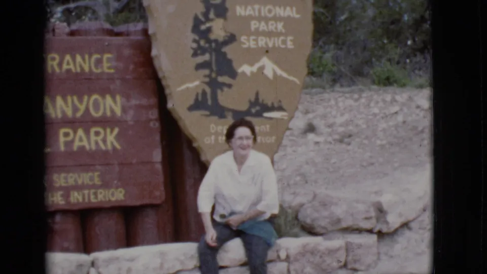 A woman in glasses smiles toward the camera beside a National Park Service entrance sign in Arizona, 1968.