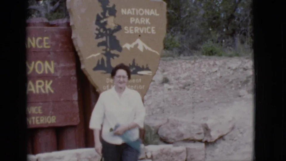 The traveler moves beside the stone ledge, with the rustic National Park Service entrance signs still visible behind her.