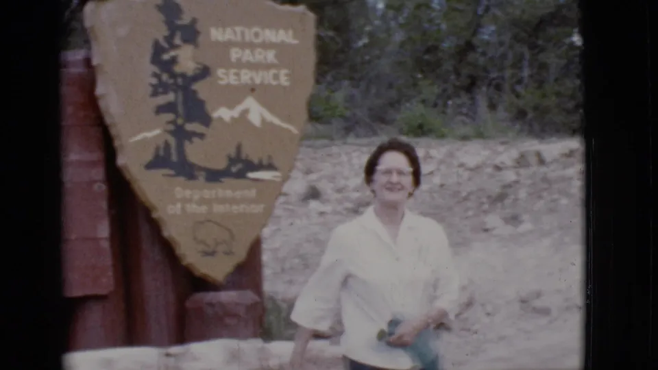 A woman in a white blouse stands by the National Park Service shield sign at an Arizona park entrance in 1968.