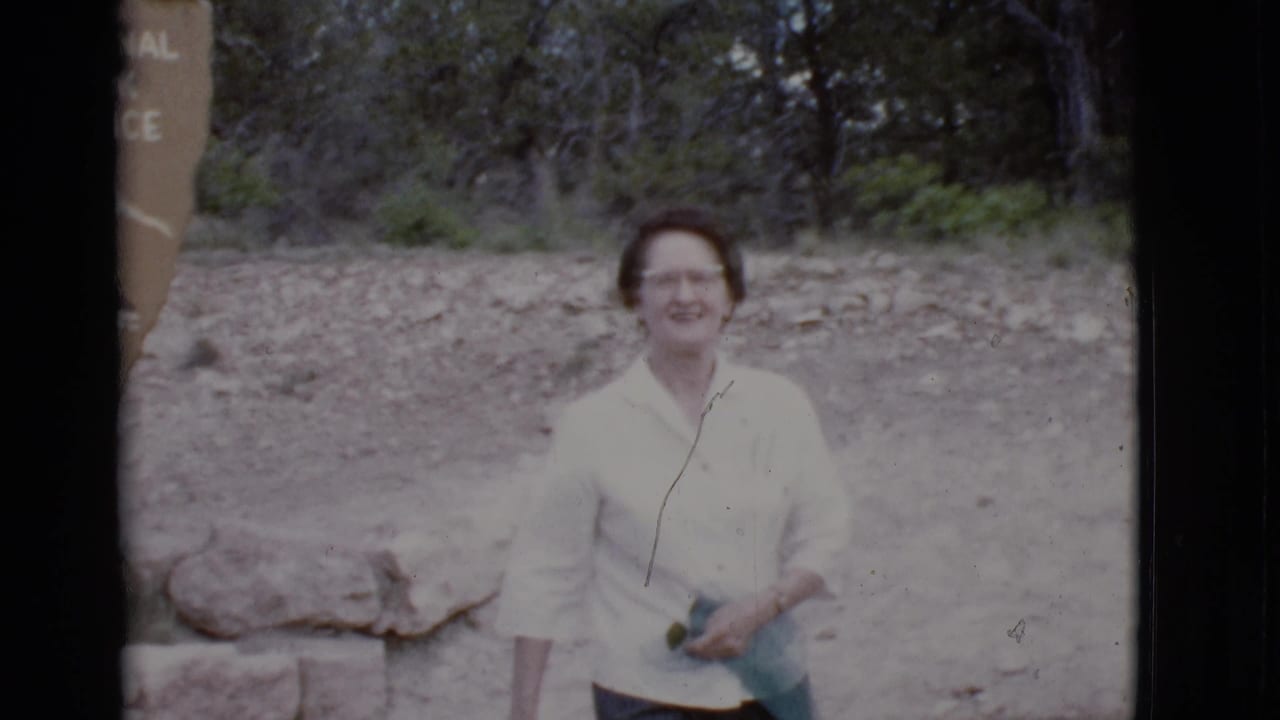 Woman in a light blouse smiles near a partial park sign on a wooded dirt path.