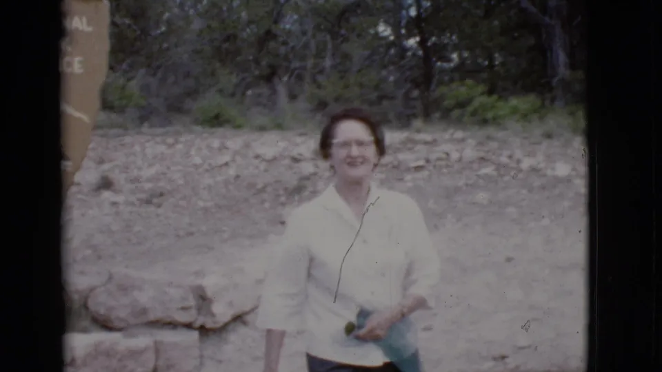 In 1968 Arizona home-movie footage, a woman in glasses smiles toward the camera beside rocks, trees, and a partial National Park Service sign.